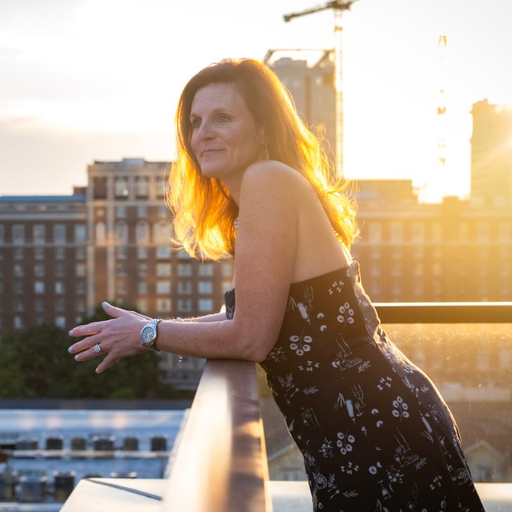 Tara Samuels in a floral dress leans on a railing at sunset, city buildings and cranes in the background.