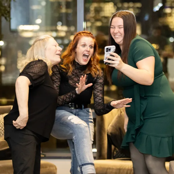 Three women smiling and laughing together while looking at a phone in a brightly lit room.