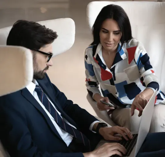 Two people in business attire discuss work and business communication while looking at a laptop; one points at the screen.