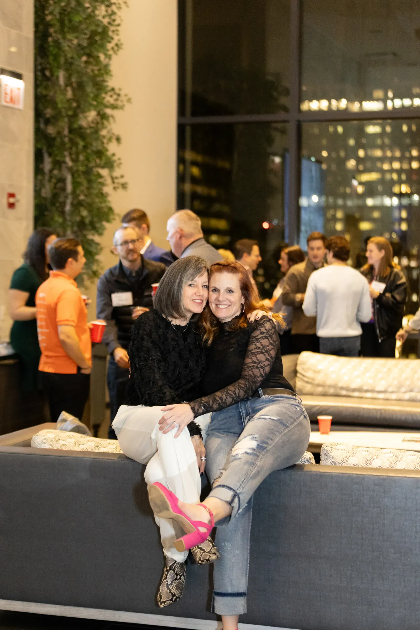 Two women sitting and smiling on a couch at a lively indoor gathering with people in the background.