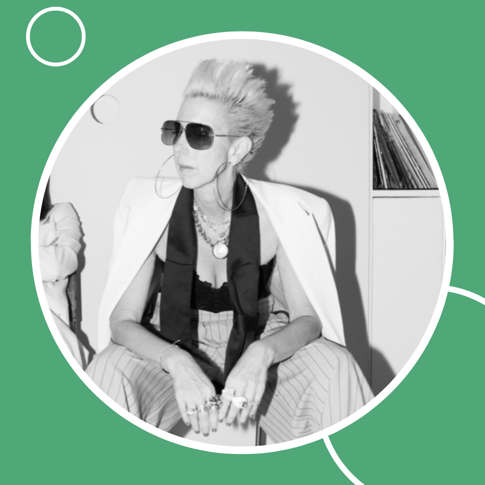 Black and white photo of a stylish person with spiked hair, sunglasses, and bold jewelry, sitting confidently.