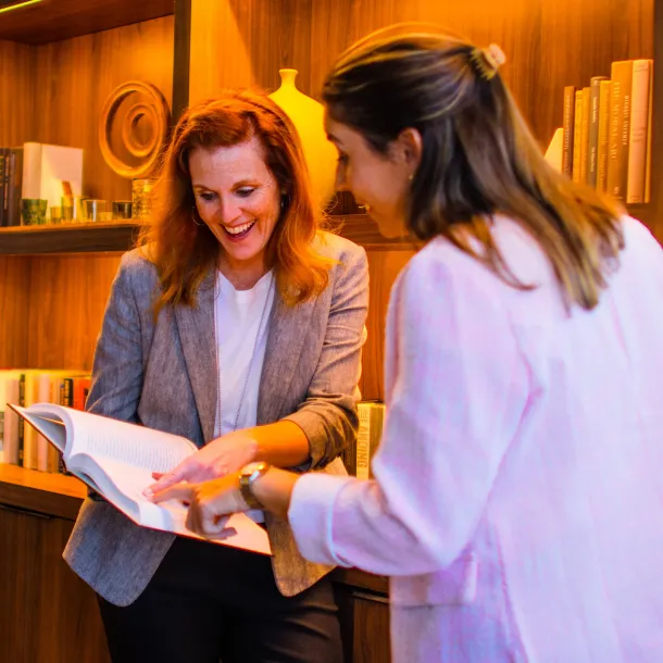 Two women smiling and looking at a book together in front of a wooden bookshelf.