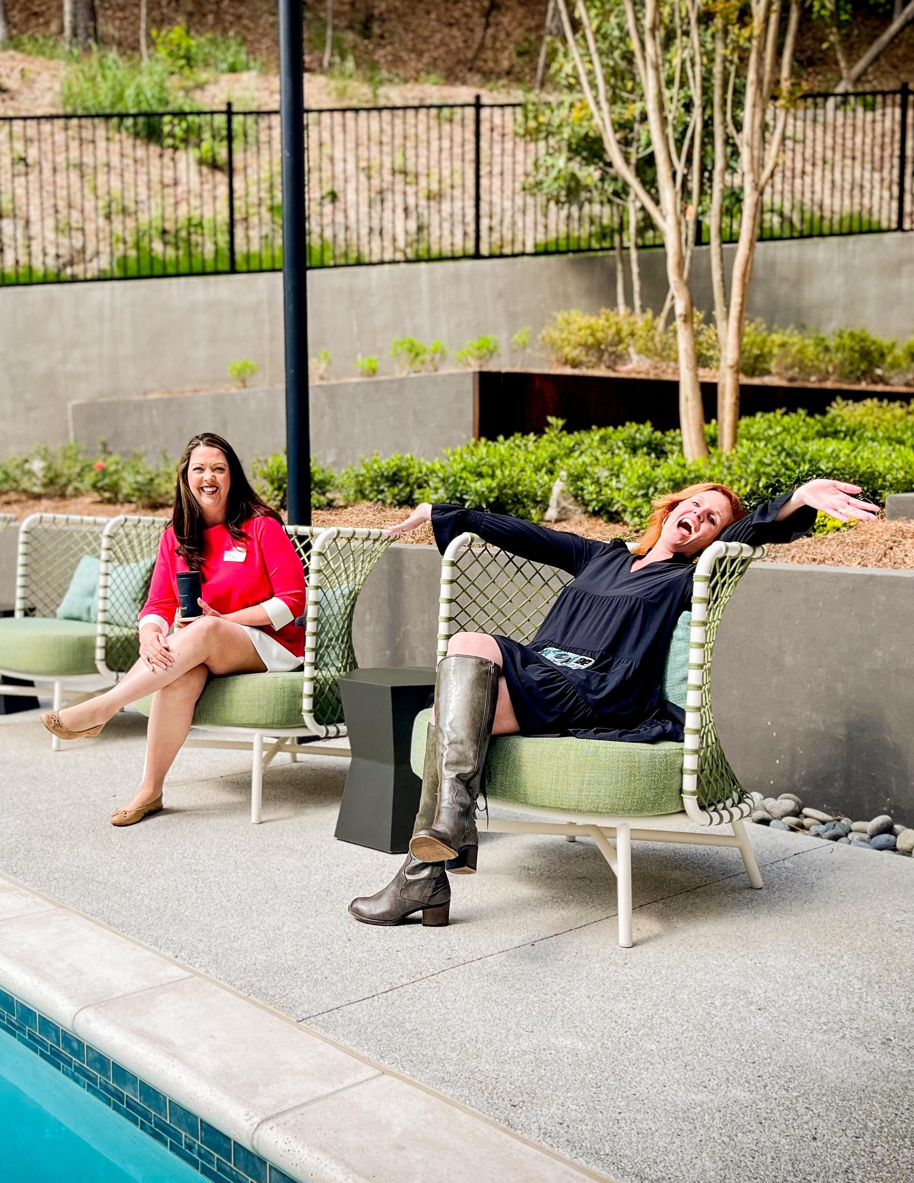 Two women sit on chairs by a pool; one sits upright, the other lounges with arms raised and smiling.